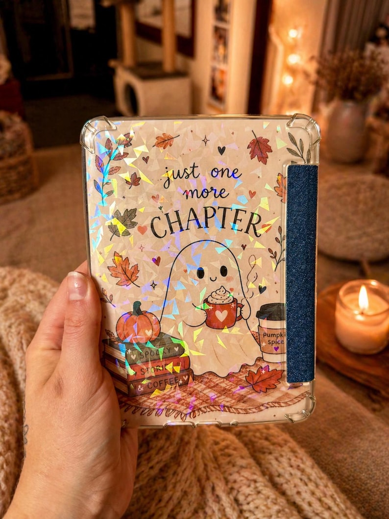 May include: A clear e-reader case featuring a holographic design of a ghost holding a red mug, a pumpkin, a stack of books labelled Spooky, Story, and Coffee, and autumn leaves. The text reads just one more CHAPTER. A dark blue textured strap is attached.