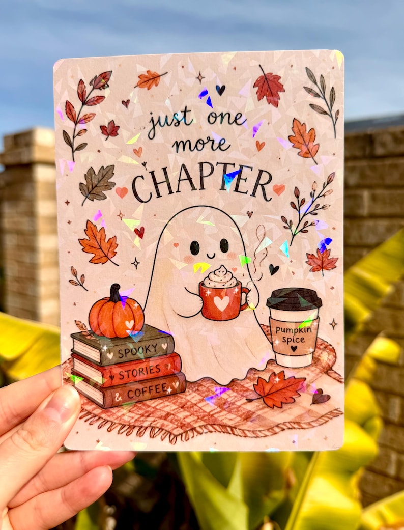May include: A rectangular holographic sticker featuring a ghost holding a red mug, a pumpkin, a stack of books labelled Spooky Stories & Coffee, and a cup labelled pumpkin spice. The text reads just one more CHAPTER amidst autumn leaves.