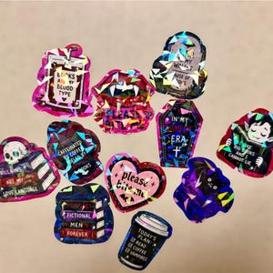 May include: A collection of holographic stickers with various designs and text. The stickers feature images of vampires, coffins, bats, and coffee cups. Text includes phrases like "Looks are my blood type" and "Villains are my love language."