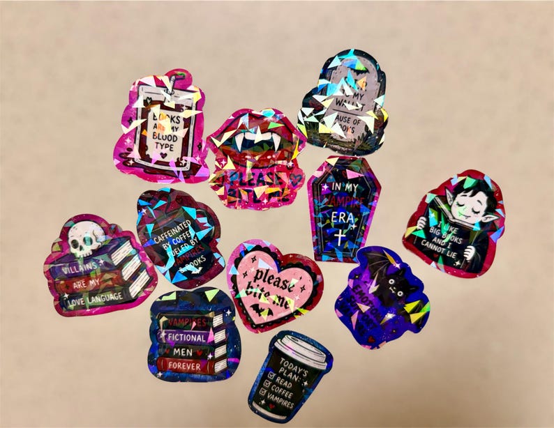 May include: A collection of holographic stickers with various designs and text. The stickers feature images of vampires, coffins, bats, and coffee cups. Text includes phrases like "Looks are my blood type" and "Villains are my love language."