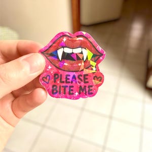 May include: A pink sticker featuring a pair of lips with vampire fangs. The lips have a glittery effect and the text "PLEASE BITE ME" is written below. The sticker has a heart on each side of the text.