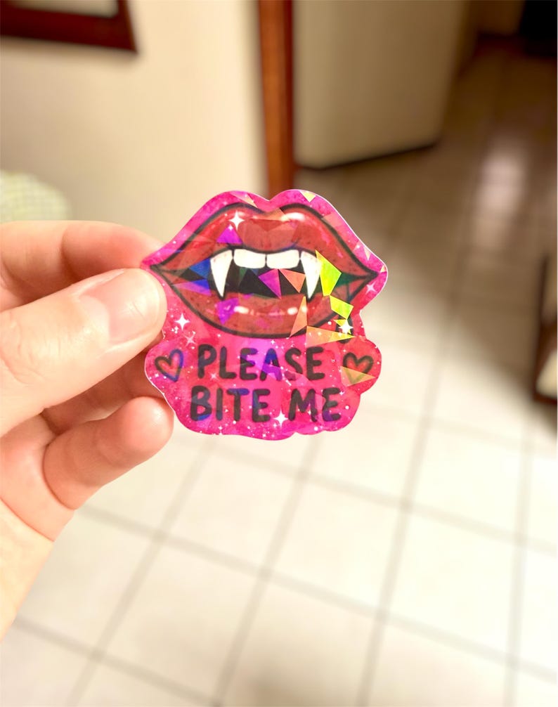May include: A pink sticker featuring a pair of lips with vampire fangs. The lips have a glittery effect and the text "PLEASE BITE ME" is written below. The sticker has a heart on each side of the text.