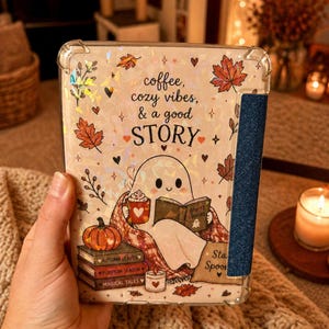 May include: A clear protective tablet case featuring a holographic autumn-themed design with a ghost reading a book, a pumpkin, and stacked books labelled Autumn Leaves, Pumpkin Season, and Magical Tales, with the text coffee, cozy vibes, & a good STORY.