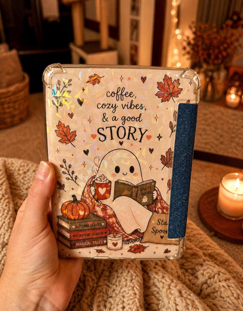 May include: A clear protective tablet case featuring a holographic autumn-themed design with a ghost reading a book, a pumpkin, and stacked books labelled Autumn Leaves, Pumpkin Season, and Magical Tales, with the text coffee, cozy vibes, & a good STORY.