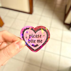 May include: A heart-shaped sticker with a pink and red design. The sticker has the words "please bite me" in a gothic font, with a bat graphic at the bottom. The sticker has a holographic, glittery finish.