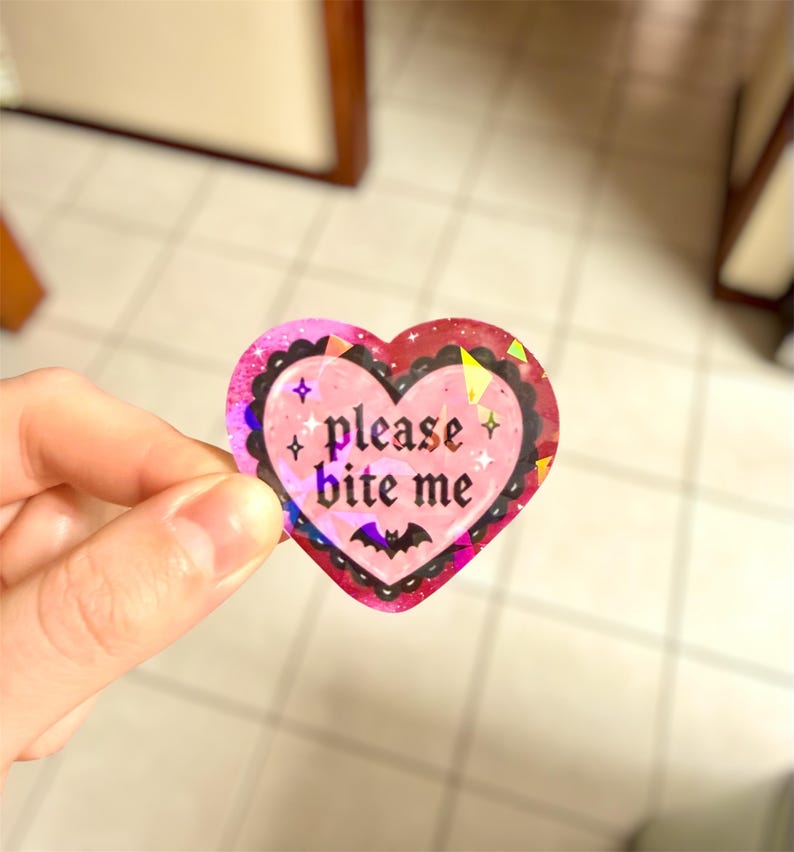 May include: A heart-shaped sticker with a pink and red design. The sticker has the words "please bite me" in a gothic font, with a bat graphic at the bottom. The sticker has a holographic, glittery finish.