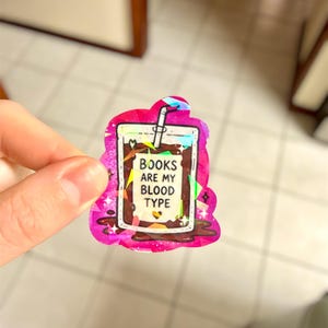May include: A holographic sticker in the shape of a drink with the text "BOOKS ARE MY BLOOD TYPE". The sticker has a pink border with a glittery effect and a white straw.