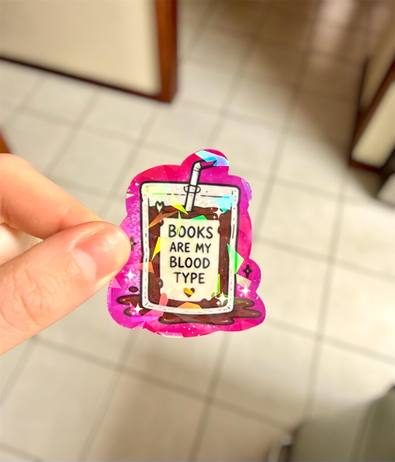 May include: A holographic sticker in the shape of a drink with the text "BOOKS ARE MY BLOOD TYPE". The sticker has a pink border with a glittery effect and a white straw.