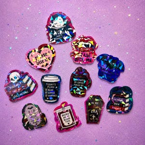 May include: A collection of holographic stickers with various designs and text. Designs include a vampire reading a book, a coffee cup, a coffin, and a heart. The stickers are in shades of pink, blue, and purple.