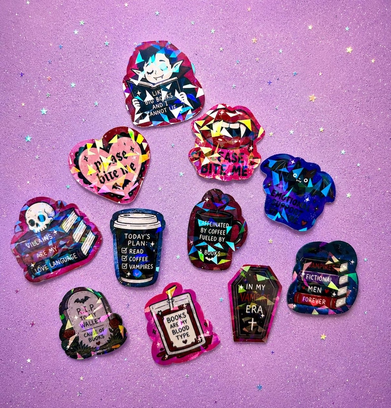May include: A collection of holographic stickers with various designs and text. Designs include a vampire reading a book, a coffee cup, a coffin, and a heart. The stickers are in shades of pink, blue, and purple.