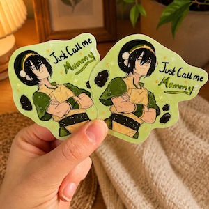 May include: Two die-cut stickers featuring an anime-style character with black hair, wearing a green and yellow outfit with a black belt. The stickers have a light green background with small black rock shapes and the text Just Call me Mommy.