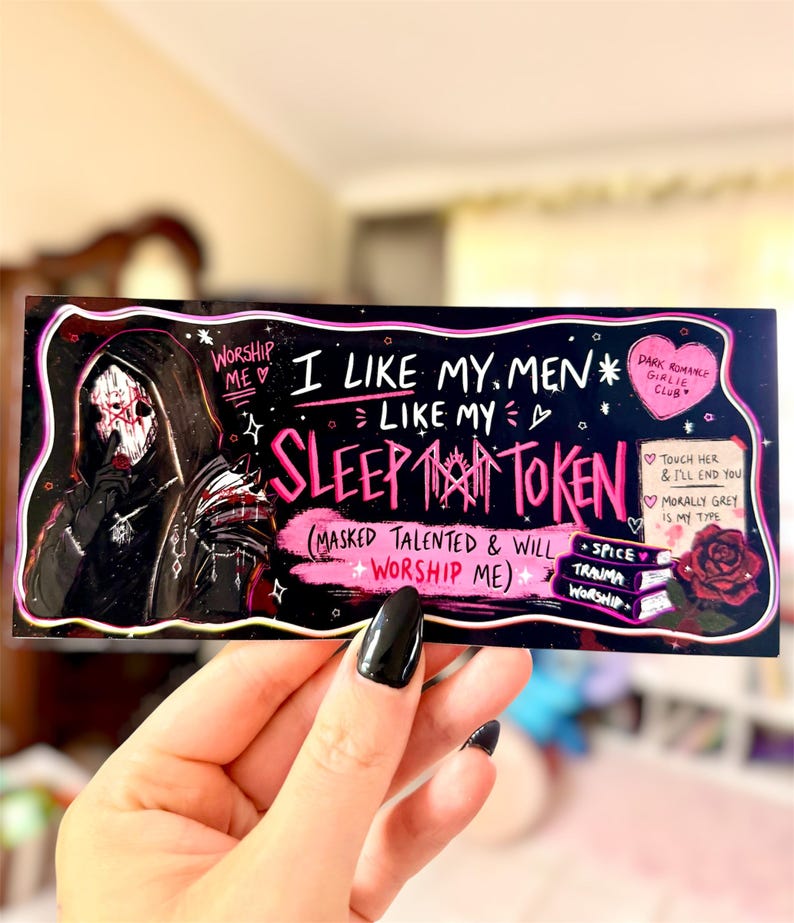 May include: A rectangular sticker with a black background and pink and white text. The sticker features an illustration of a figure in a hooded cloak and the text "I LIKE MY MEN LIKE MY SLEEP TOKEN". Additional text includes "Worship Me" and "Dark Romance Girlie Club".