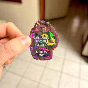 May include: A holographic sticker in the shape of a coffee mug. The sticker has the text "Caffeinated by coffee, fueled by vampires & books." The mug is dark with colorful geometric shapes and a bat graphic.