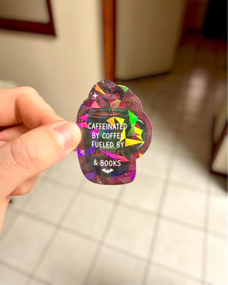 May include: A holographic sticker in the shape of a coffee mug. The sticker has the text "Caffeinated by coffee, fueled by vampires & books." The mug is dark with colorful geometric shapes and a bat graphic.