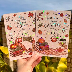 May include: Two rectangular holographic prints featuring a ghost character. The left print says "coffee, cozy vibes, & a good STORY" with autumn leaves, a pumpkin, and books. The right print says "just one more CHAPTER" with a ghost, pumpkin, and coffee.