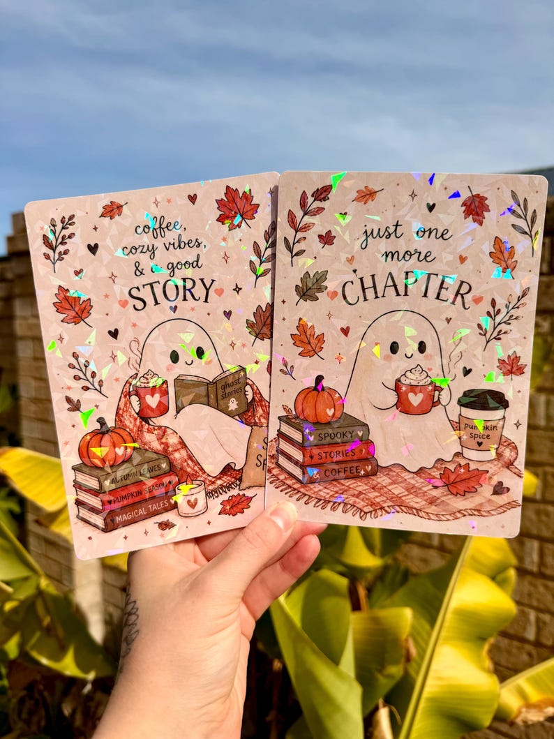 May include: Two rectangular holographic prints featuring a ghost character. The left print says "coffee, cozy vibes, & a good STORY" with autumn leaves, a pumpkin, and books. The right print says "just one more CHAPTER" with a ghost, pumpkin, and coffee.