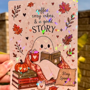 May include: A rectangular holographic sticker featuring a ghost reading a book titled ghost stories, holding a red mug with a heart. The text reads coffee, cozy vibes, & a good STORY and Stay Spooky. Includes autumn leaves, a pumpkin, and books.
