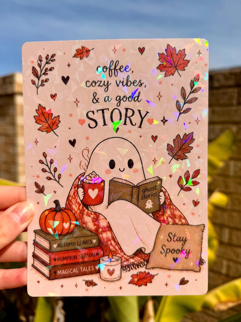 May include: A rectangular holographic sticker featuring a ghost reading a book titled ghost stories, holding a red mug with a heart. The text reads coffee, cozy vibes, & a good STORY and Stay Spooky. Includes autumn leaves, a pumpkin, and books.