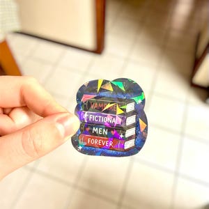 May include: A holographic sticker featuring a stack of four books. The books are labeled "VAMPIRE", "FICTIONAL", "MEN", and "FOREVER". The sticker has a dark blue background with a starry design and geometric shapes.