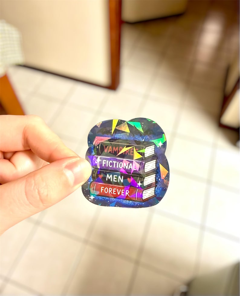 May include: A holographic sticker featuring a stack of four books. The books are labeled "VAMPIRE", "FICTIONAL", "MEN", and "FOREVER". The sticker has a dark blue background with a starry design and geometric shapes.
