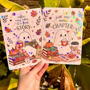 May include: Two rectangular holographic prints featuring a ghost character. The left print says coffee, cozy vibes, & a good STORY with a ghost reading a book. The right print says just one more CHAPTER with a ghost holding a mug and a pumpkin.