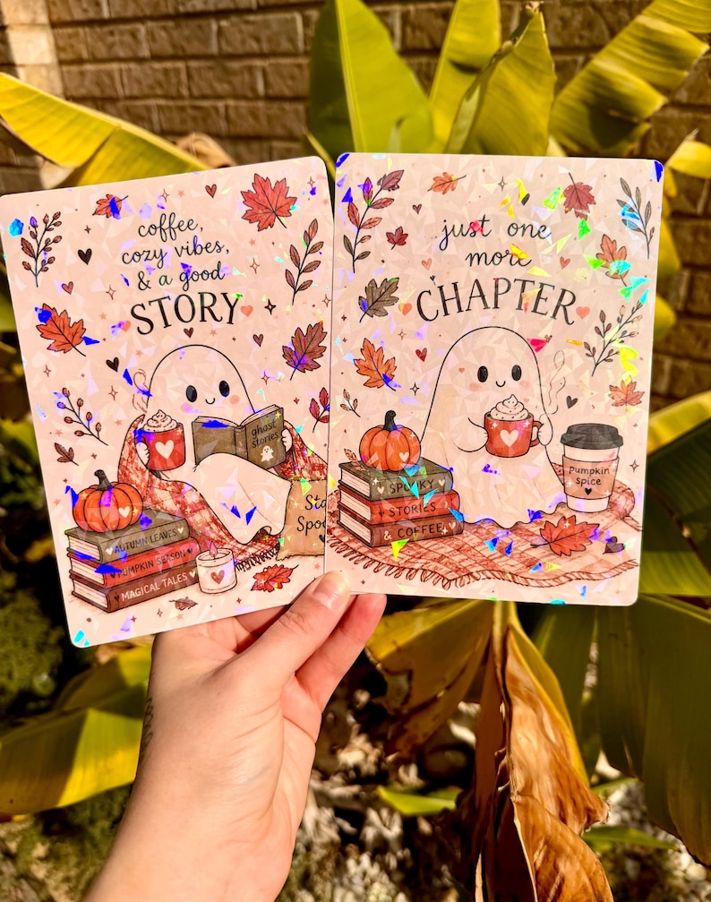 May include: Two rectangular holographic prints featuring a ghost character. The left print says coffee, cozy vibes, & a good STORY with a ghost reading a book. The right print says just one more CHAPTER with a ghost holding a mug and a pumpkin.