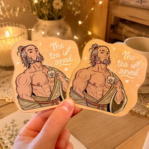 May include: Two die-cut stickers featuring an illustration of a muscular, bearded character with a topknot, wearing a robe and holding a small teacup. The text "The tea is served" is printed in white script alongside small star and heart icons.