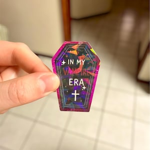 May include: A holographic coffin-shaped sticker with the text "IN MY VAMPIRE ERA" and a cross symbol. The sticker has a pink and purple border and a geometric design. The sticker is held by a hand.