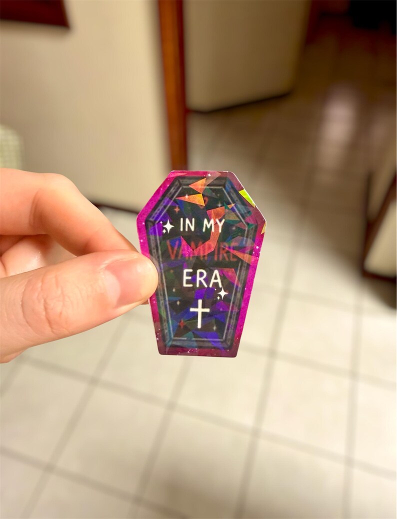 May include: A holographic coffin-shaped sticker with the text "IN MY VAMPIRE ERA" and a cross symbol. The sticker has a pink and purple border and a geometric design. The sticker is held by a hand.