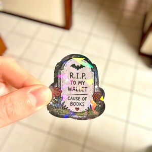 May include: A holographic sticker in the shape of a tombstone. The sticker reads "R.I.P. TO MY WALLET CAUSE OF BOOKS" with a bat and roses. The sticker is held by a hand.