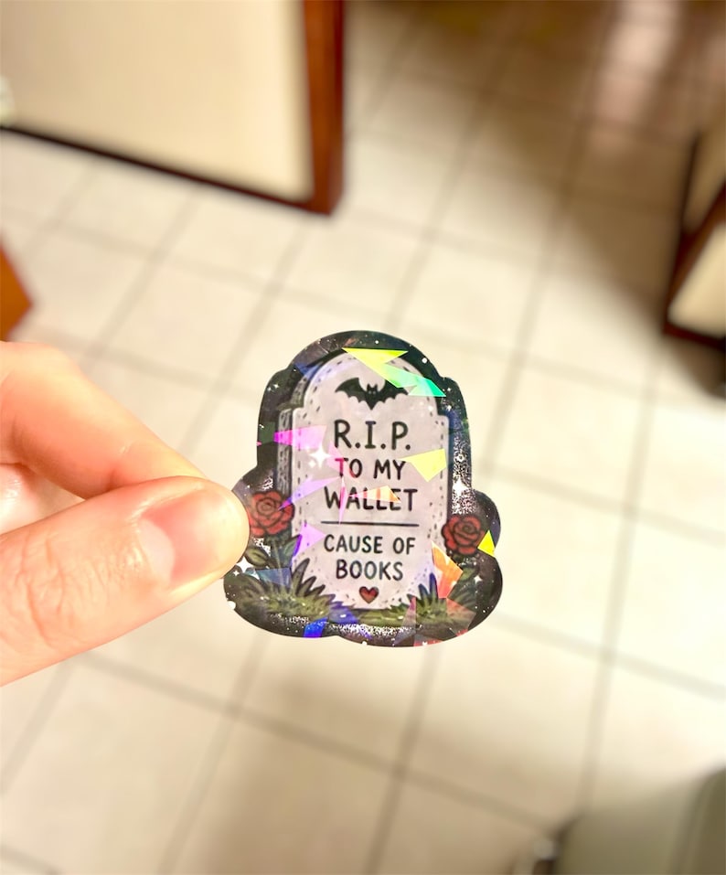 May include: A holographic sticker in the shape of a tombstone. The sticker reads "R.I.P. TO MY WALLET CAUSE OF BOOKS" with a bat and roses. The sticker is held by a hand.
