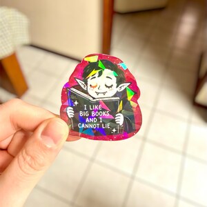 May include: A holographic sticker featuring a cartoon vampire reading a book. The vampire has closed eyes and pointed ears. The book reads "I LIKE BIG BOOKS AND I CANNOT LIE." The sticker has a pink and black border with iridescent accents.