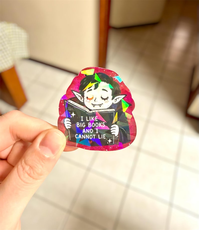 May include: A holographic sticker featuring a cartoon vampire reading a book. The vampire has closed eyes and pointed ears. The book reads "I LIKE BIG BOOKS AND I CANNOT LIE." The sticker has a pink and black border with iridescent accents.