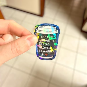 May include: A holographic sticker in the shape of a coffee cup. The sticker has a blue background with the text "TODAY'S PLAN:" followed by check boxes for "READ", "COFFEE", and "VAMPIRES". The sticker has a glittery, iridescent finish.