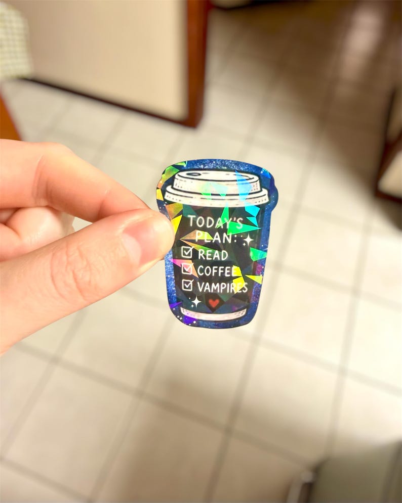 May include: A holographic sticker in the shape of a coffee cup. The sticker has a blue background with the text "TODAY'S PLAN:" followed by check boxes for "READ", "COFFEE", and "VAMPIRES". The sticker has a glittery, iridescent finish.