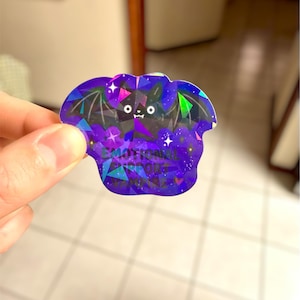 May include: A holographic sticker in the shape of a bat, featuring a geometric design in shades of purple, blue, and green. The sticker has the words "EMOTIONAL SUPPORT" printed on it. The bat has a cute face and wings.