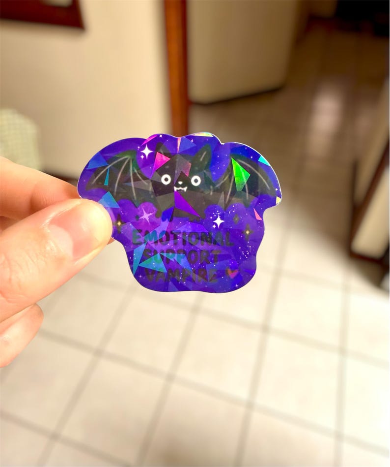 May include: A holographic sticker in the shape of a bat, featuring a geometric design in shades of purple, blue, and green. The sticker has the words "EMOTIONAL SUPPORT" printed on it. The bat has a cute face and wings.