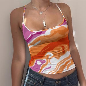 May include: A colorful tank top with a vibrant abstract pattern in shades of pink, orange, and white. The top has thin spaghetti straps and a scoop neckline. The bottom half of the image shows a pair of blue denim jeans.