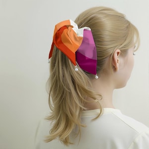 May include: A colorful hair bow featuring shades of orange, red, white, and purple. The bow is adorned with small, white pearl accents at the tips. The hair accessory is tied in a ponytail.