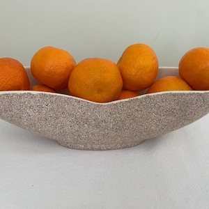 May include: A decorative, oblong bowl filled with several bright orange tangerines. The bowl has a speckled, textured exterior in shades of beige and brown. The tangerines are round and have a slightly rough texture.