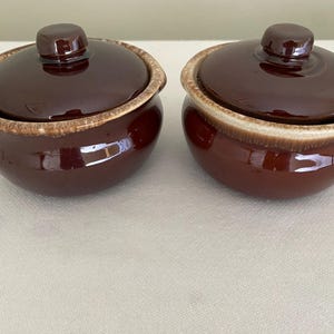 May include: Two matching brown ceramic bowls with lids. Each bowl has a rounded shape with a lid and a light brown rim. The bowls are glossy and appear to be vintage kitchenware.