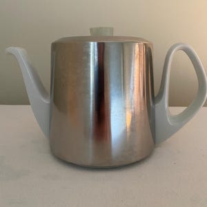 May include: A stainless steel teapot with a white handle, spout, and lid accents. The teapot has a rounded body and a curved handle. The lid has a small, light-colored knob. The teapot is designed for brewing tea.