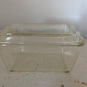 May include: A clear, rectangular glass baking dish with a matching lid. The dish has slightly flared sides and a decorative edge on the lid. The glass has a slight yellow tint, suggesting its age. The dish is sitting on a distressed white surface.