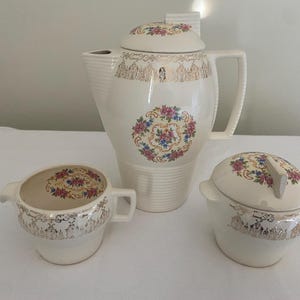 May include: A cream-colored vintage tea set with floral and gold accents. The set includes a teapot, creamer, and sugar bowl. The teapot has a tall, slender design with a lid, and the creamer and sugar bowl have matching floral patterns.