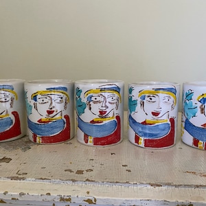 May include: Set of five white ceramic mugs with colorful hand-painted designs of faces. Each mug features a different face with blue, red, yellow, and green accents.