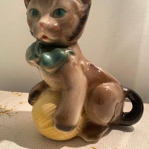 May include: A brown ceramic cat figurine with blue eyes and a teal bow. The cat is sitting on a yellow ball of yarn.