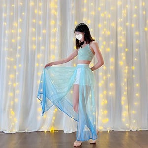May include: A woman wearing a light blue and white ombre sheer skirt with a white top and white shorts. The skirt is flowing and has a long train. The woman is standing in front of a white curtain with string lights.