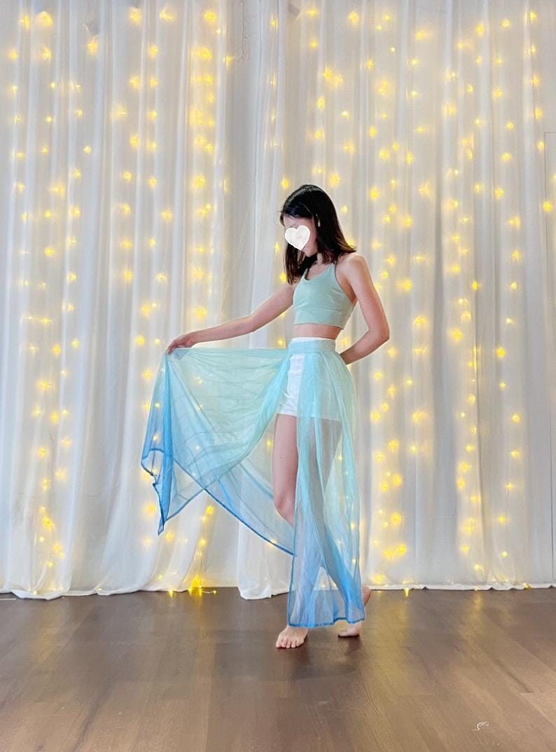 May include: A woman wearing a light blue and white ombre sheer skirt with a white top and white shorts. The skirt is flowing and has a long train. The woman is standing in front of a white curtain with string lights.
