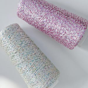 May include: Two spools of decorative thread. One spool is a light purple color with iridescent sequins, and the other is a light silver color with iridescent sequins. The spools are cylindrical and have clear plastic bases.