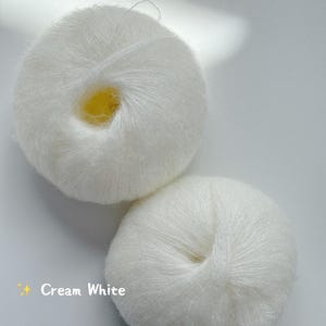 Puede incluir: Dos ovillos de hilo blanco crema se muestran sobre un fondo blanco. El hilo parece suave y esponjoso, con un ligero brillo. El texto "Cream White" es visible en la imagen, indicando el color del hilo.